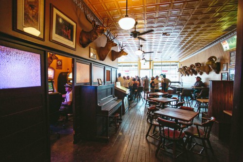 A rustic bar interior with wooden tables, a piano, and mounted animal heads, bustling with patrons enjoying their time.