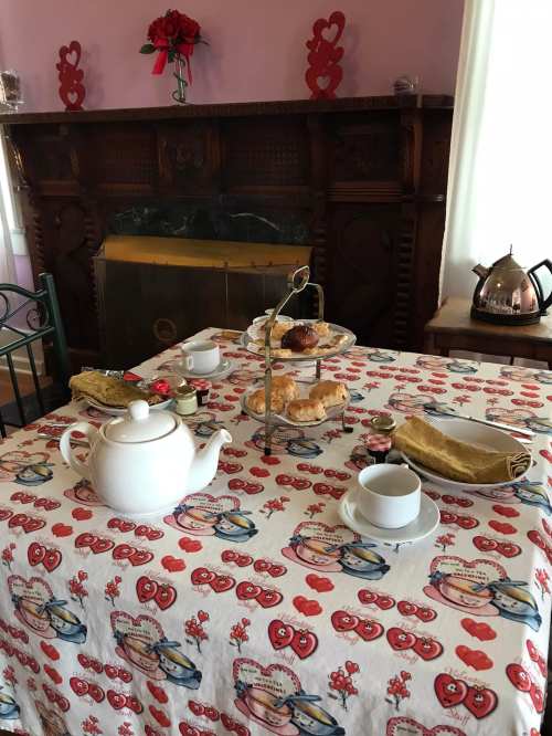 A cozy tea setting with a white teapot, cups, pastries, and a decorated tablecloth in a charming room.