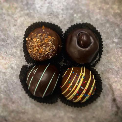 Four assorted chocolate truffles in black paper cups, topped with nuts and colorful drizzles.