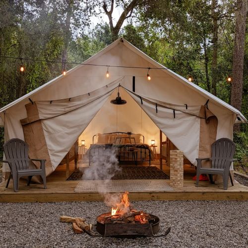 A cozy glamping tent with a bed, chairs, and a fire pit outside, surrounded by trees and string lights.