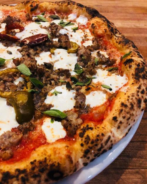 Close-up of a pizza topped with sausage, pepperoni, mozzarella, jalapeños, and fresh herbs on a charred crust.