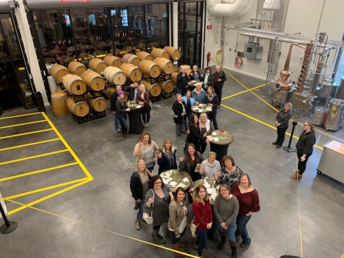 A group of people gathered in a spacious distillery, with barrels and equipment in the background.