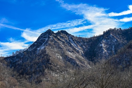 A rugged mountain peak with a snowy summit under a bright blue sky and wispy clouds.