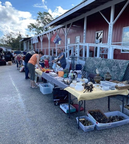 A bustling outdoor flea market with vendors selling various antiques and collectibles along a gravel path.