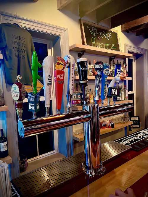 A bar with various beer taps, including IPAs and stouts, against a backdrop of shelves and a welcome sign.