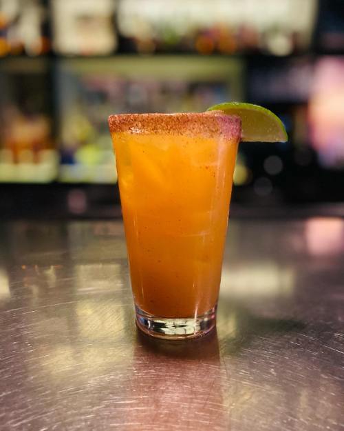 A tall glass of orange cocktail with a salted rim and a lime wedge, set on a bar counter.