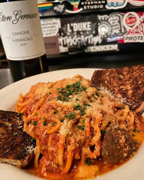 A plate of spaghetti with meatballs, topped with cheese and herbs, alongside toasted bread and a bottle of red wine.