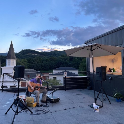 A musician plays guitar on a rooftop stage with mountains and a cloudy sky in the background.