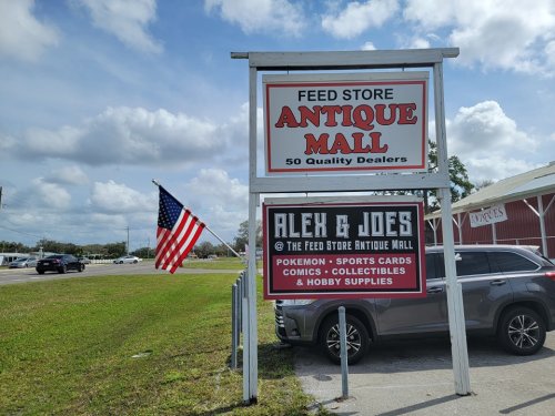 Sign for "Feed Store Antique Mall" with "Alex & Joe's" below, featuring an American flag and grassy area.