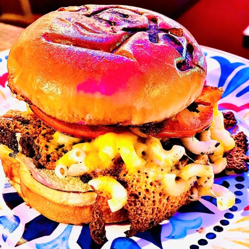 A close-up of a burger with a toasted bun, melted cheese, macaroni, and bacon on a colorful plate.