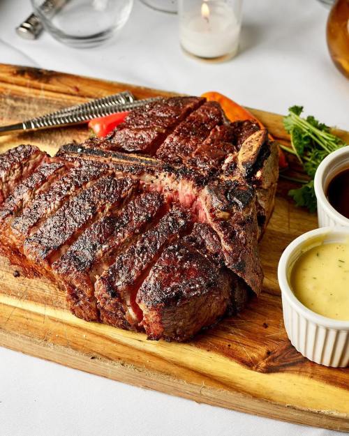 A perfectly cooked steak on a wooden board, accompanied by two sauces and garnished with herbs.