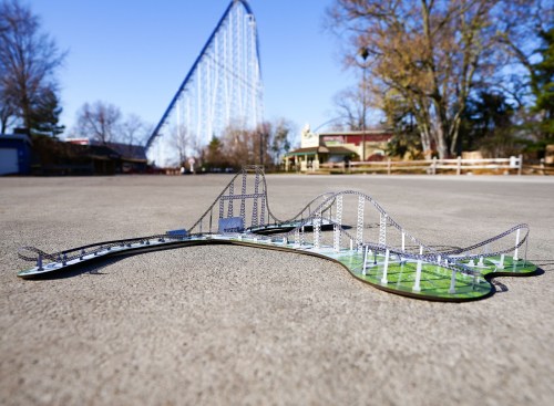 A miniature roller coaster model on a flat surface, with a large roller coaster in the background.