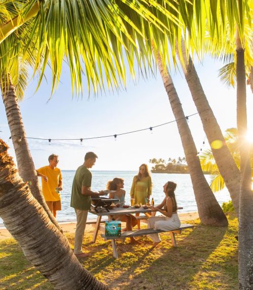 A group of friends enjoying a barbecue by the beach, surrounded by palm trees and sunset lighting.