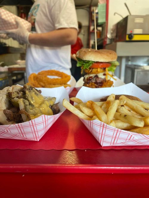 A close-up of a table with a burger, fries, and a cheesy meat dish, with a person in the background preparing food.