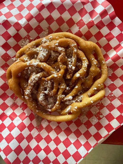 A golden-brown funnel cake dusted with powdered sugar on a red and white checkered paper.