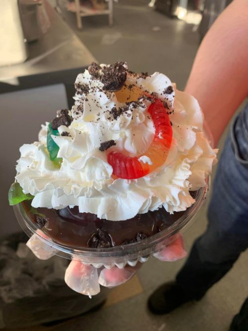 A hand holding a dessert cup topped with whipped cream, crushed cookies, and gummy worms.