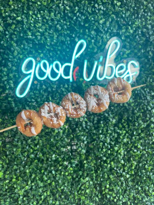 A skewer of mini donuts drizzled with icing, set against a green backdrop with a neon "good vibes" sign.