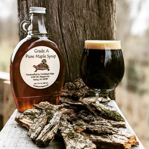 A bottle of Grade A pure maple syrup next to a glass of dark beer, with bark pieces on a wooden surface outdoors.