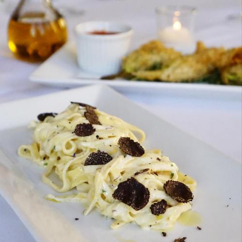 Plate of creamy pasta topped with black truffle shavings, garnished with herbs, served in an elegant dining setting.