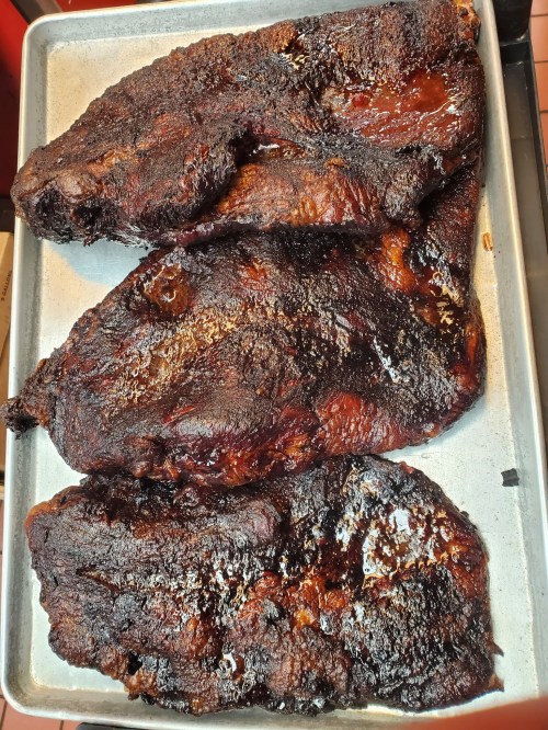 Three large, smoked meat cuts on a metal tray, with a dark, charred exterior and a glossy finish.