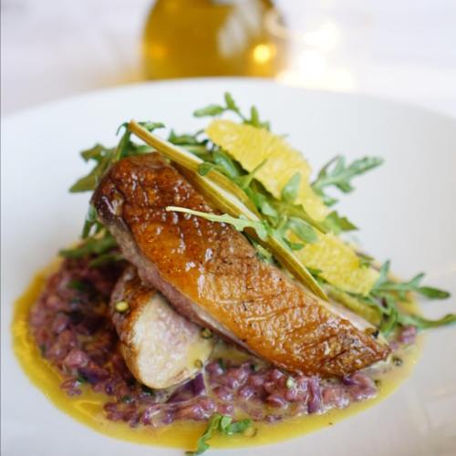 A beautifully plated dish featuring seared duck breast on a bed of purple rice, garnished with greens and citrus.