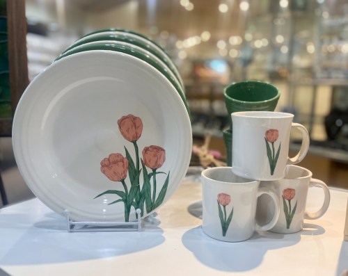 A set of white dishes and mugs featuring pink tulip designs, displayed on a table with a blurred background.