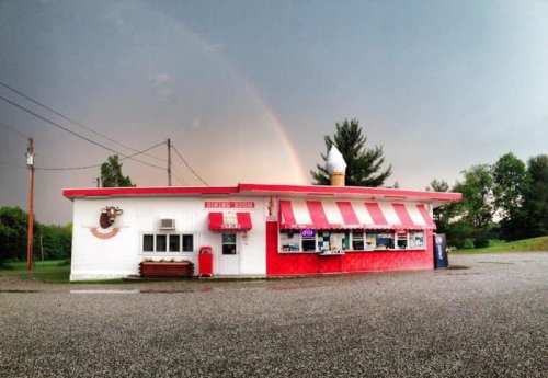 A colorful ice cream shop with a red and white striped awning, set against a backdrop of a rainbow and cloudy sky.