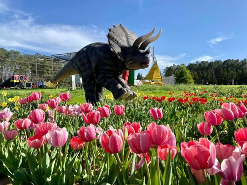 A dinosaur statue stands among colorful tulips in a sunny field, with trees and a pyramid structure in the background.