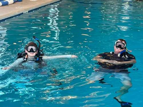 Two people in scuba gear swimming in a pool, surrounded by clear blue water.