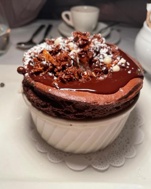 A rich chocolate soufflé topped with chocolate sauce and crunchy bits, served in a white ramekin.