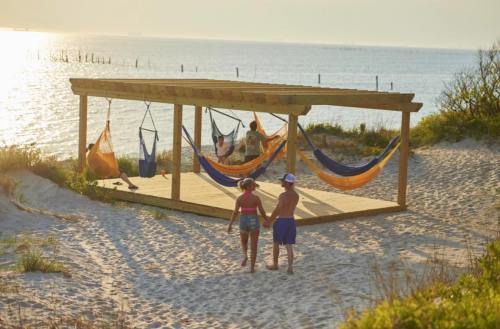 Two children walk hand in hand on a sandy beach near a wooden structure with colorful hammocks at sunset.