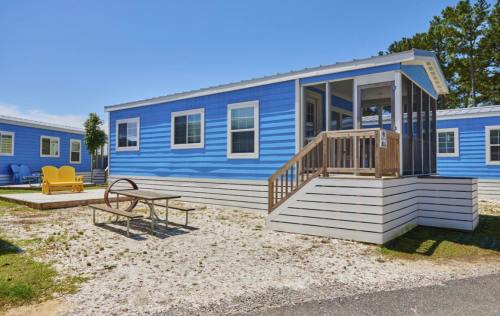 A blue mobile home with a wooden porch and steps, surrounded by gravel and green grass, under a clear blue sky.