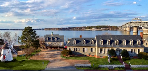 Scenic view of a waterfront with historic buildings, a windmill, and a bridge under a partly cloudy sky.