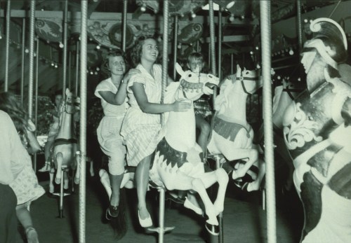 Two girls joyfully riding white carousel horses in a vintage amusement park setting.