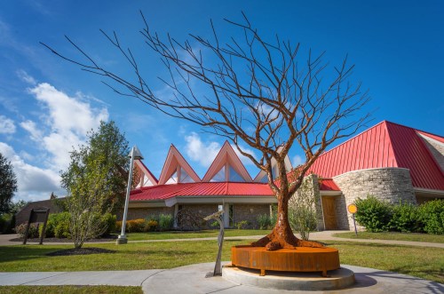 A large, bare tree sculpture stands in front of a modern building with a red roof and triangular peaks under a blue sky.