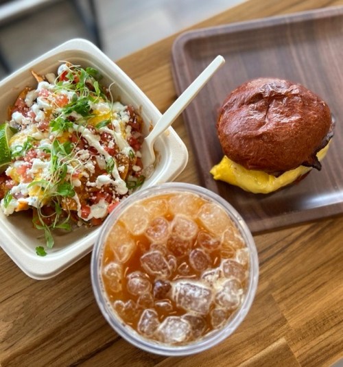 A bowl of nachos with toppings, a sandwich on a wooden plate, and a cup of iced tea on a wooden table.