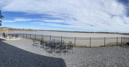 Panoramic view of a river with a fenced patio area and scattered chairs under a blue sky with clouds.