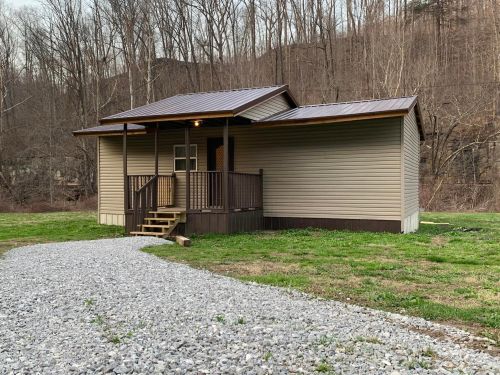 A small, single-story house with a porch, surrounded by trees and a gravel path in a rural setting.