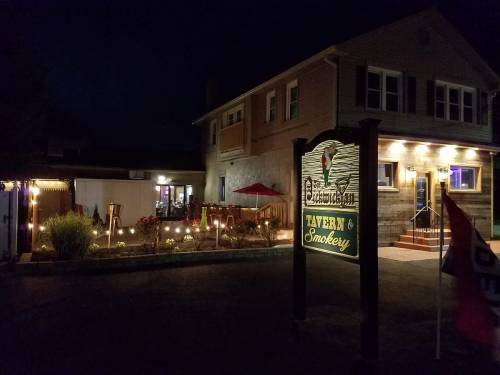 A nighttime view of a tavern and smokery with a welcoming sign, outdoor seating, and soft lighting.
