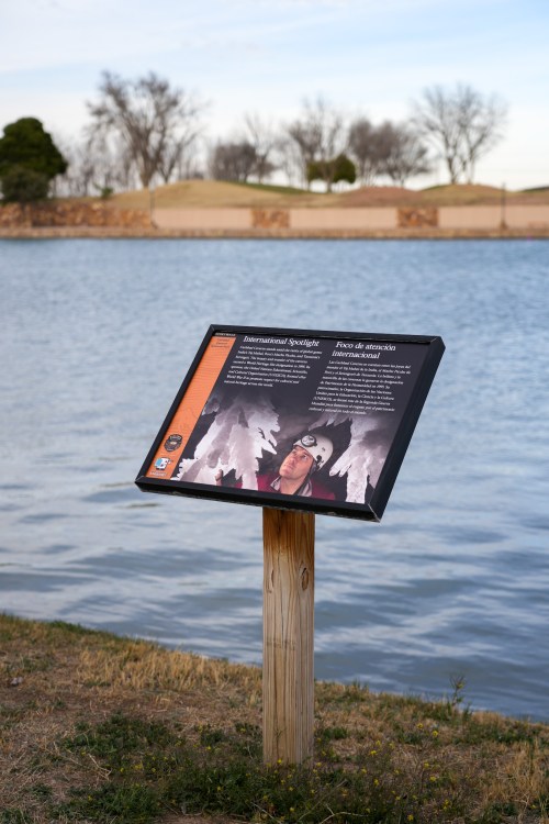 A sign by a lake, featuring information about speleology, with a person in a helmet and cave imagery.
