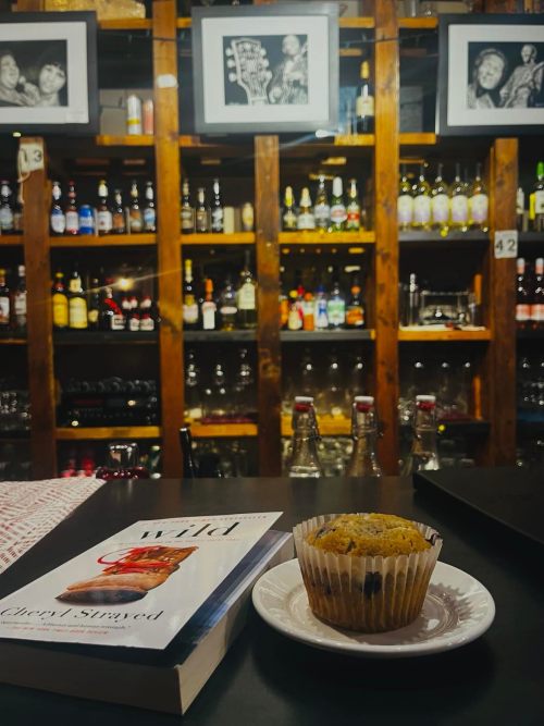 A cozy bar scene featuring a muffin on a plate, a book titled "Wild," and shelves lined with bottles and framed photos.