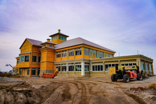 A large, partially constructed building with orange sheathing and a gravel path, featuring construction equipment nearby.