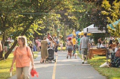 A bustling outdoor market scene with people walking, shopping, and enjoying the day under trees and tents.