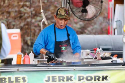 A man in a blue shirt and camo hat cooks jerk pork at a food stall, surrounded by condiments and smoke.