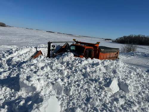 A snowplow is partially buried in deep snow, surrounded by a vast snowy landscape under a clear blue sky.