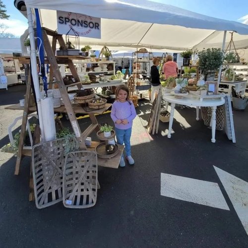 A young girl stands in front of a market stall filled with plants and decorative items under a tent.