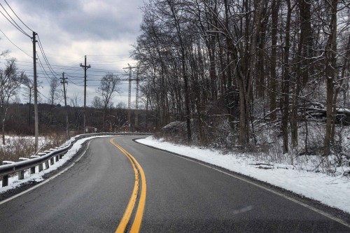 A winding road lined with snow and trees, under a cloudy sky, with power lines in the background.