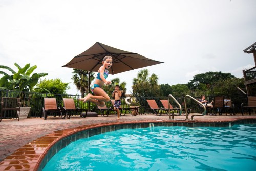 A child jumps into a pool while another child watches nearby, surrounded by lounge chairs and tropical plants.