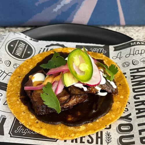 A taco topped with grilled meat, onions, jalapeños, cilantro, and sour cream on a decorative paper background.