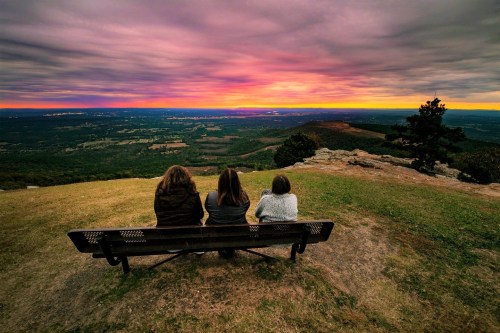 Three people sit on a bench overlooking a scenic landscape at sunset, with vibrant colors in the sky.